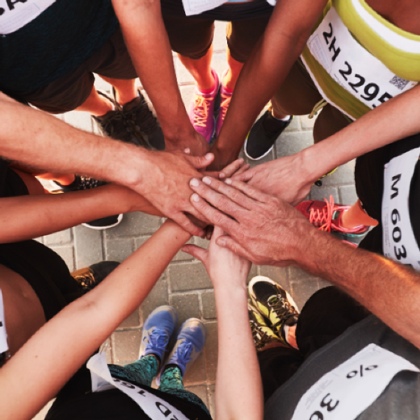 Hands of a group of runner coming together in a circle, with running shoes and pavement visible below